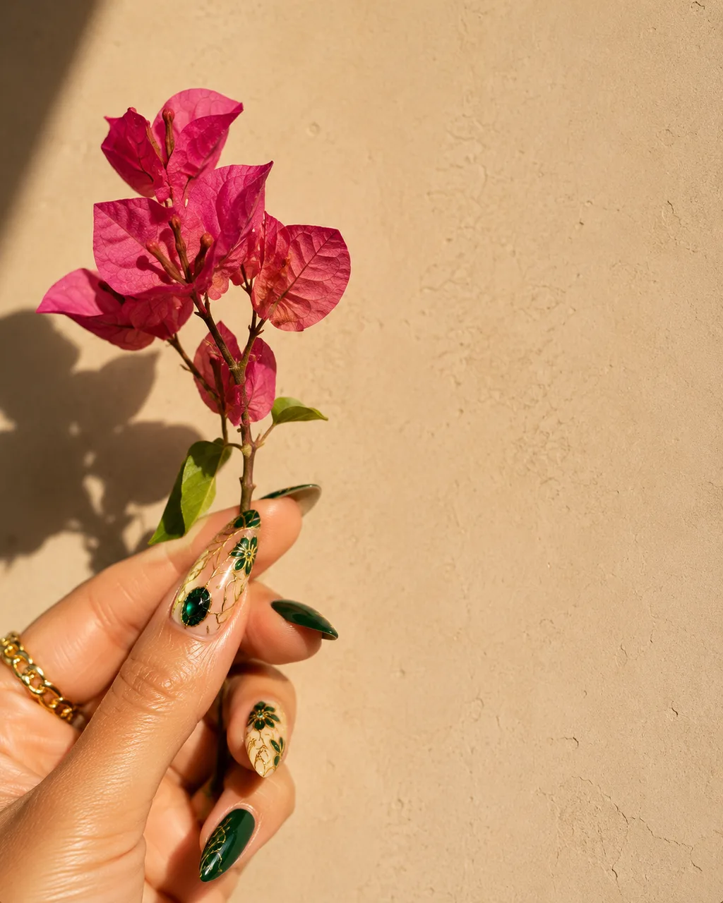 Hand-painted nail art with emerald and gold accents holding a pink bougainvillea sprig